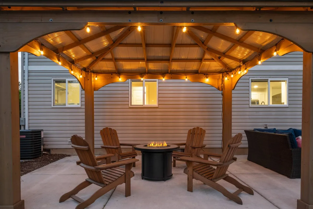 An atmospheric exterior featuring a gazebo draped in string lights, surrounded by Adirondack chairs and a crackling fire pit. The space is clearly designed for intimate gatherings and memorable evenings outdoors.