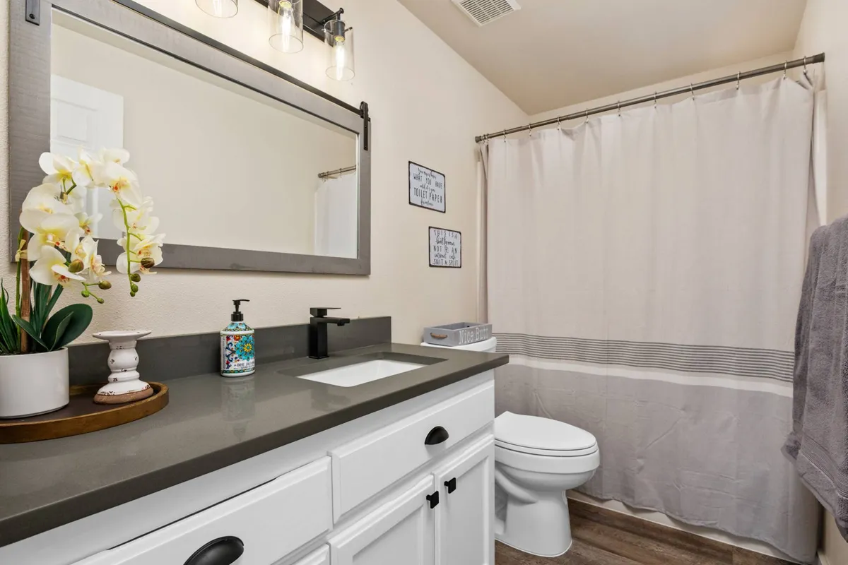 A modern, inviting bathroom with a sink featuring a contemporary faucet, a large mirror, decorative flowers, and a softly lit ambiance. The shower curtain and light fixture complete this fresh and relaxing space.