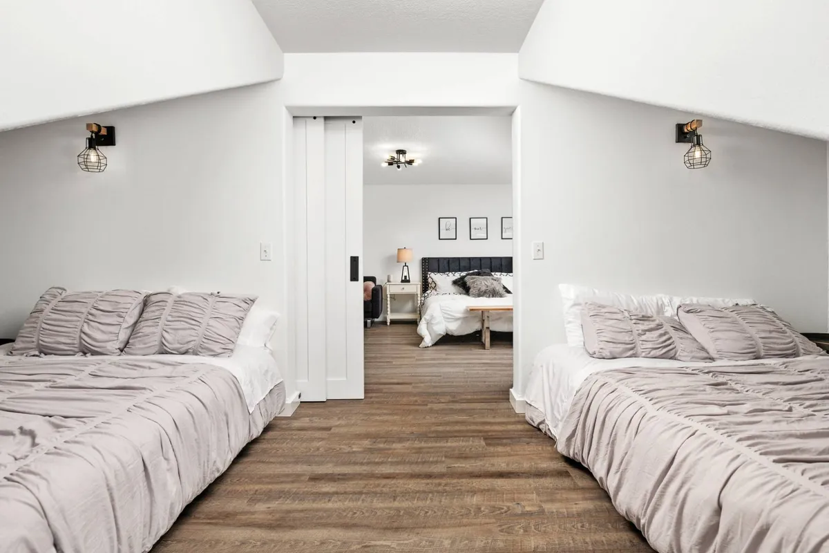 A flexible sleeping space showing two beds with soft bedding and a sliding door leading to an adjoining room, enhanced by decorative wall lights that add warmth. Sunlight streams in, highlighting the versatility of the sleeping arrangements.