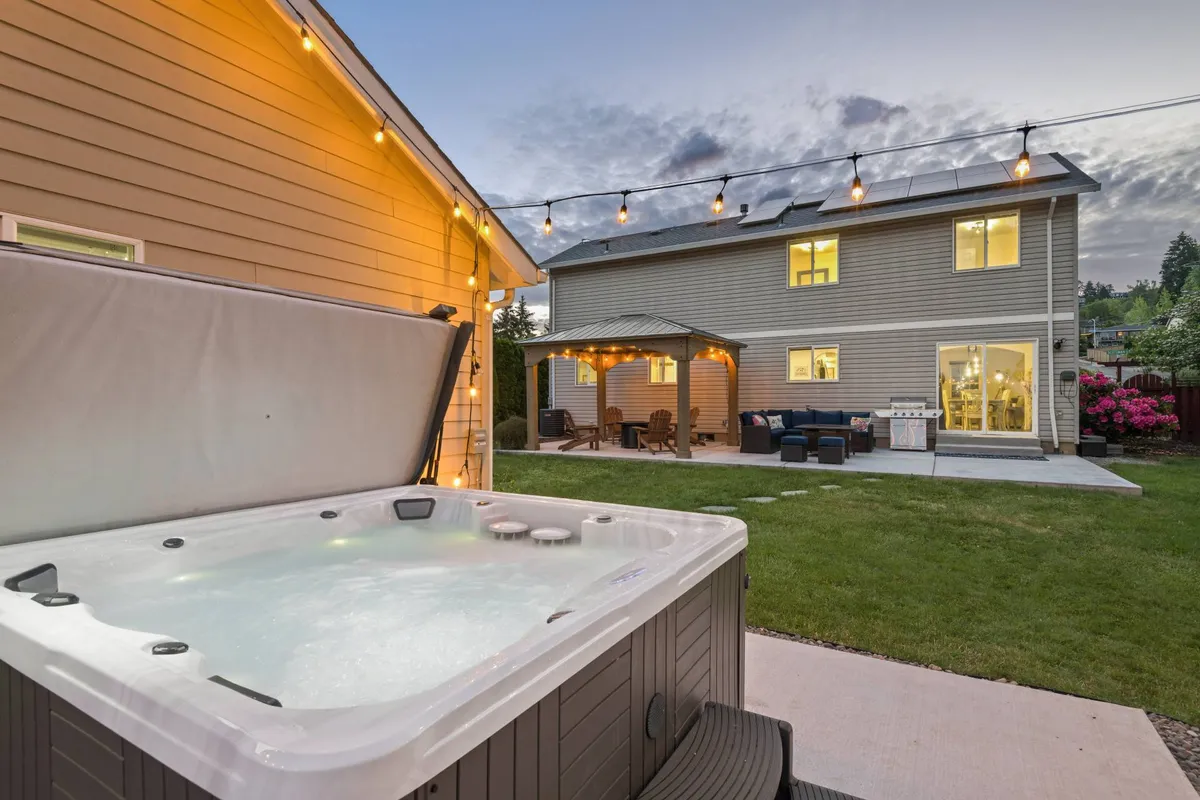 A cozy outdoor scene centered around a hot tub under hanging string lights, with plush seating nearby and the house softly illuminated in the background. The inviting setup encourages relaxation and socializing, day or night.