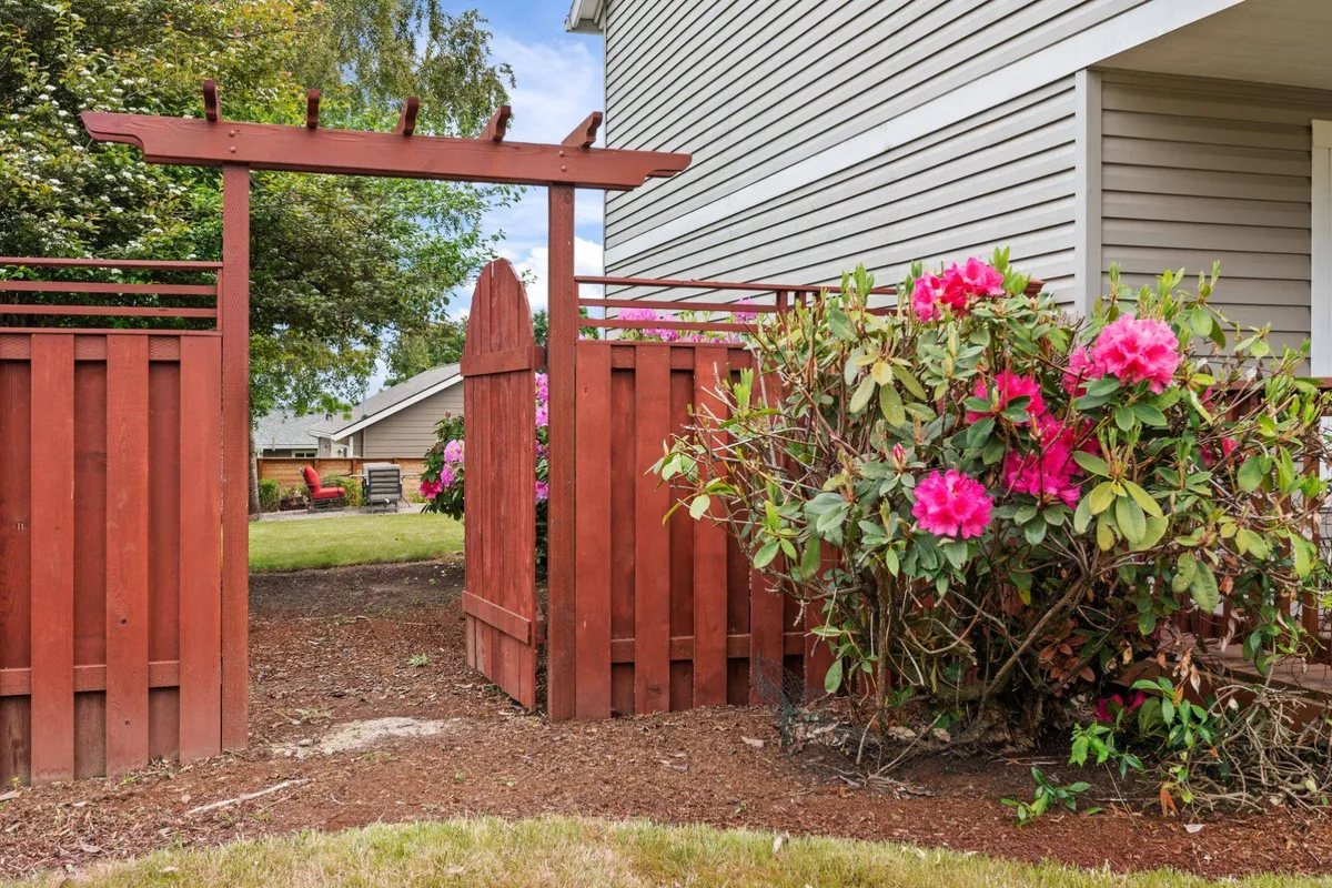 A welcoming red wooden gate framed by vibrant pink flowers and lush green shrubs, inviting guests into a serene outdoor space that connects the home to its natural surroundings.