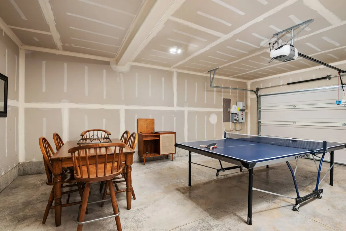 A bright and functional garage space with a ping pong table taking center stage, surrounded by wooden chairs and a dining table. The open garage door enhances the casual, welcoming atmosphere, making the space ideal for both active play and casual family gatherings.