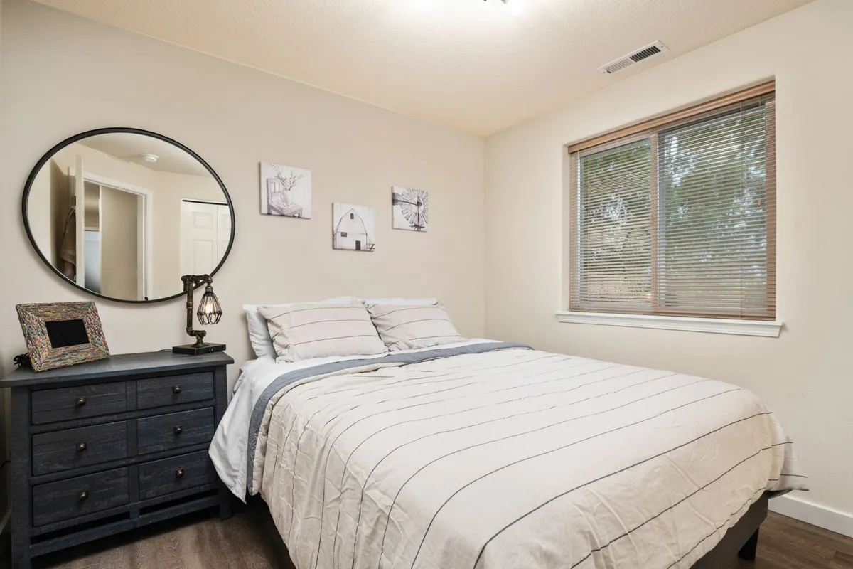 A tranquil bedroom scene featuring a king-sized bed with a nightstand, a decorative mirror, and artwork adorning the walls. Natural light filters in softly, contributing to an atmosphere of relaxation and rest.