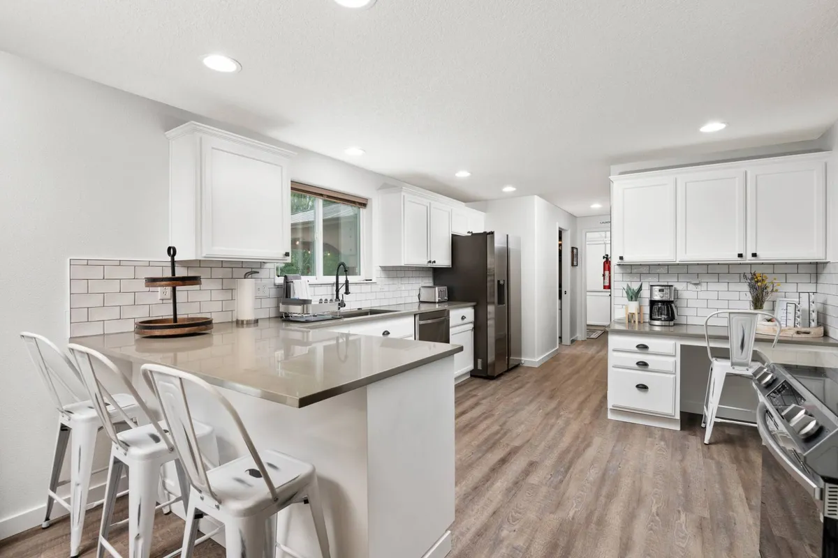 A stylish, modern kitchen featuring a large island countertop, bar stools for casual seating, sleek stainless steel appliances, and crisp white cabinetry. The kitchen is open to the living and dining space, with ample natural light and a layout perfect for both cooking and socializing.
