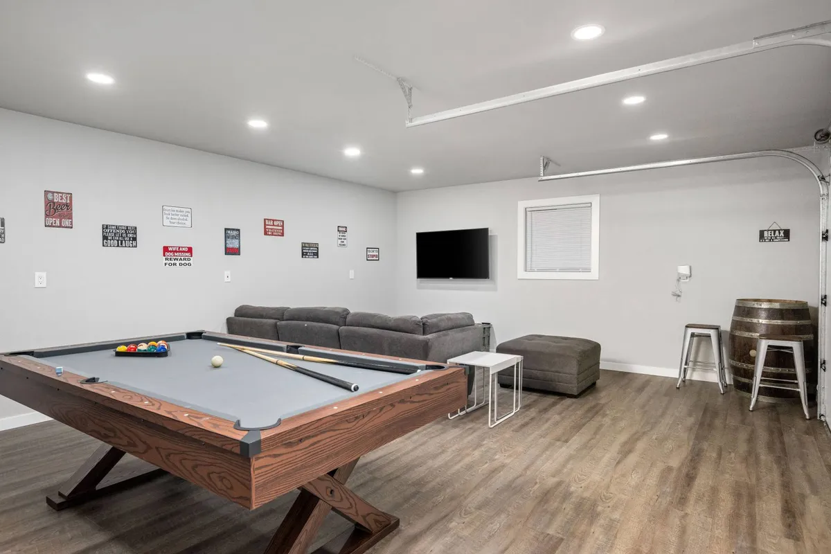 An inviting interior view of the detached garage transformed into a stylish entertainment lounge, featuring a pool table at the center, a comfortable sofa, a TV mounted on the wall, and a barrel table perfect for drinks or snacks. The space is set up for socializing and friendly competition, echoing the room's multipurpose use for games and gatherings.