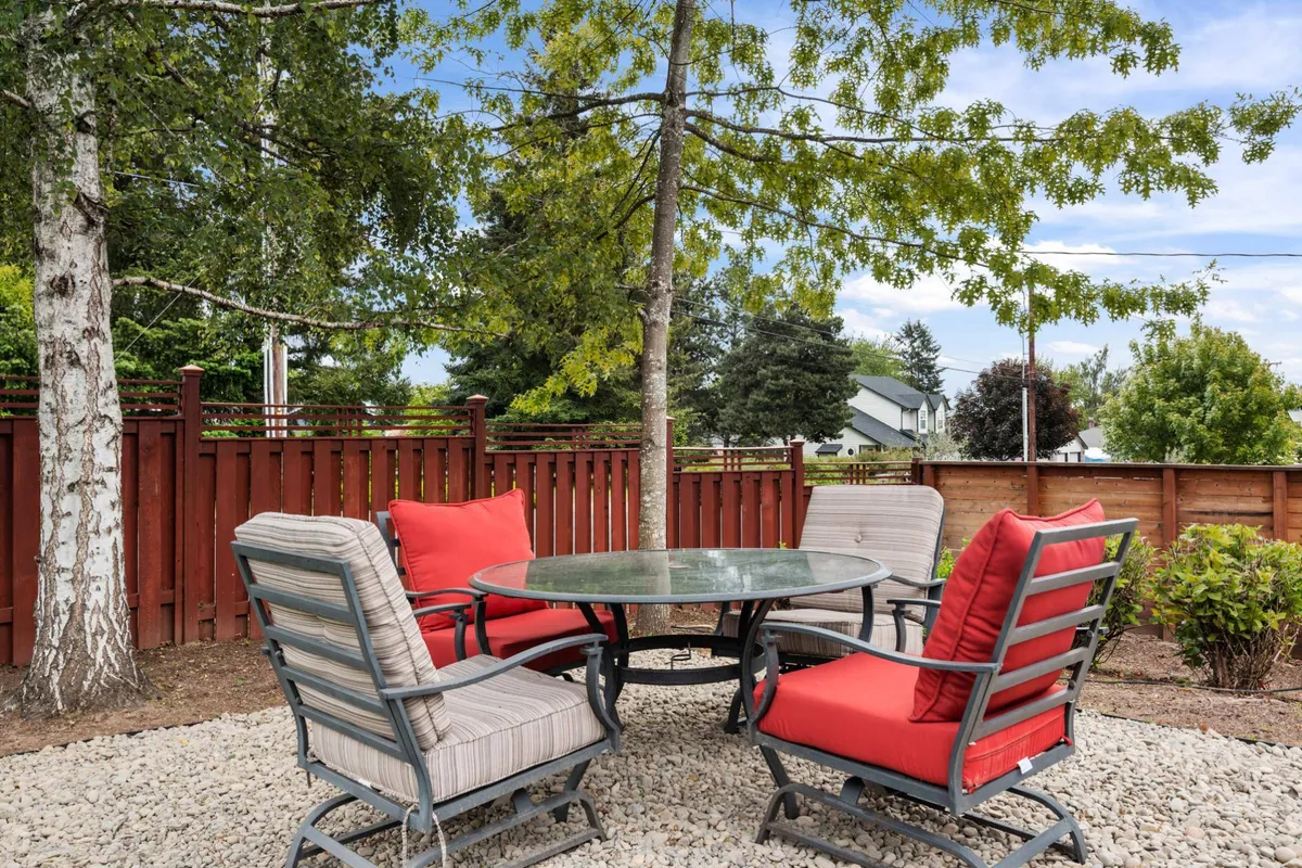 An outdoor seating arrangement featuring a round glass table with red cushions surrounded by lush green trees. The setting is ideal for outdoor dining, relaxed conversations, and soaking in the natural surroundings, with an inviting and friendly vibe.