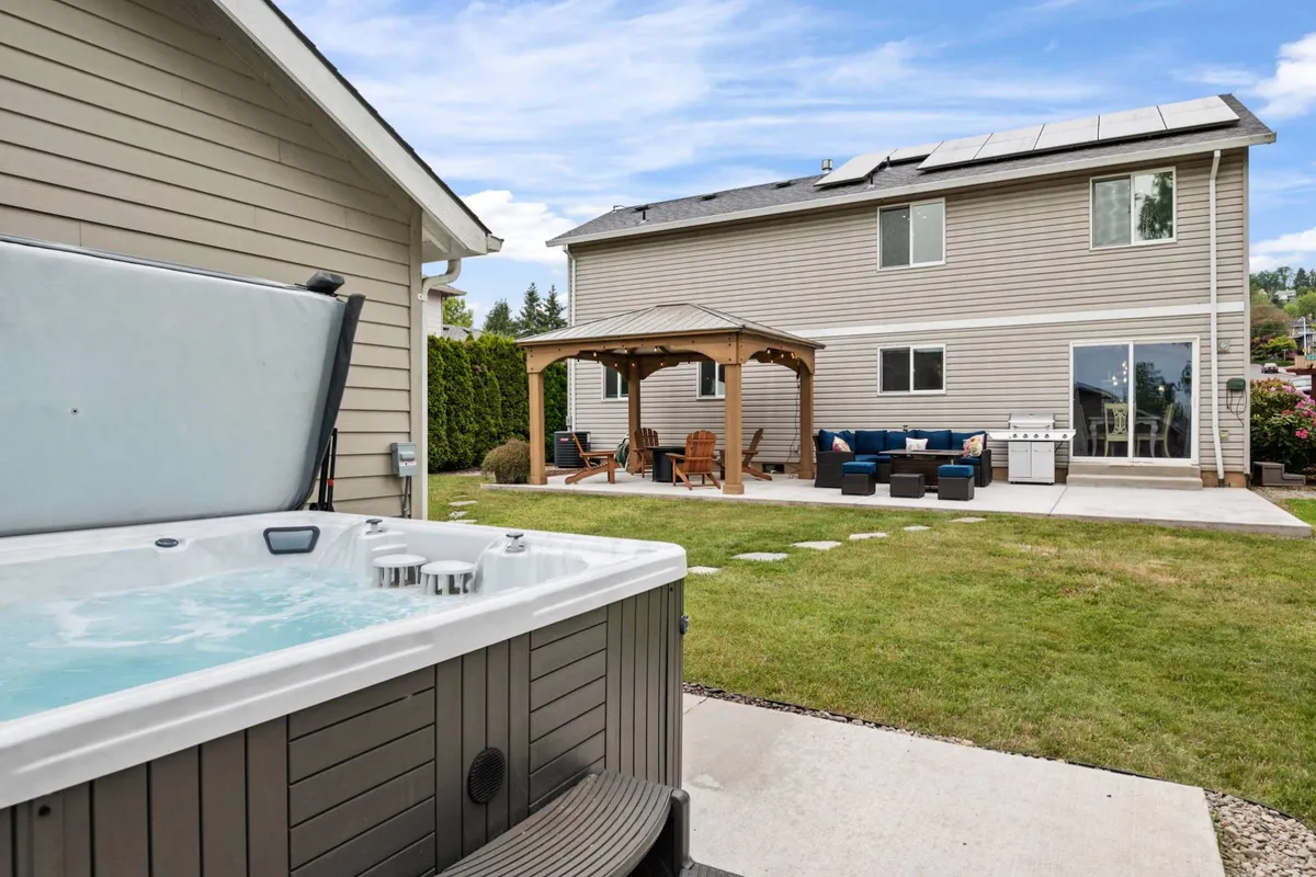 An inviting backyard scene featuring a spacious hot tub, a comfortable patio area with seating, a gazebo, and a lush lawn—the ideal setting for both family relaxation and social gatherings. The backdrop of trees and well-maintained landscaping enhances the tranquil yet playful atmosphere.