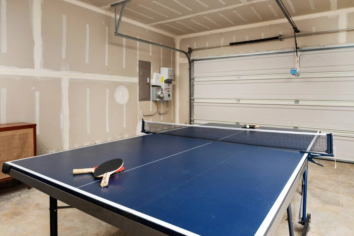A bright, playful garage lounge equipped with a ping pong table, paddles, and a raised garage door, suggesting a casual, engaging space for friendly competition and active fun. The scene is set for both kids and adults to enjoy spirited games and social time together.